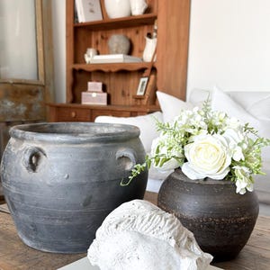 May include: A still life arrangement featuring a large, dark grey ceramic pot with handles, a small dark vase with white flowers, and a white plaster bust on a wooden table. A wooden cabinet is in the background.