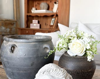 May include: A rustic still life arrangement featuring a large, dark gray ceramic pot with handles, a small dark vase with white flowers, and a white plaster bust on a wooden table. A wooden cabinet is in the background.