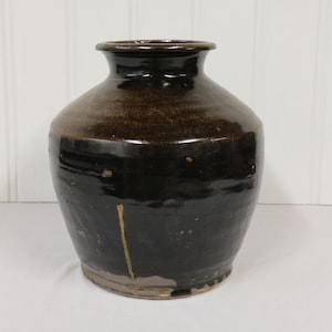 Peut inclure: Un vase en céramique brun foncé avec une finition brillante. Le vase a un corps arrondi et un col étroit. Une ligne verticale jaune descend sur le côté. Le vase est probablement à des fins décoratives.