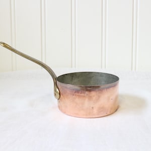 May include: A round copper saucepan with a metallic finish and a long, curved brass handle attached to the side. The interior of the pan has a silver-toned lining, and the exterior shows a warm, reddish-brown copper hue with visible surface texture.