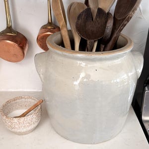 May include: A selection of wooden spoons in shades of brown and tan are stored in a large, off-white ceramic crock. A small, speckled bowl with a wooden spoon is placed nearby. Copper pots hang from a copper bar.