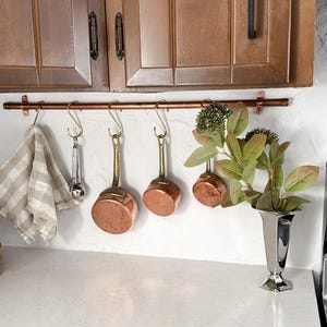 May include: A kitchen setting with copper pans and a ladle suspended from a copper rail. A checkered tea towel hangs nearby. A silver vase with foliage sits on the counter. Wooden cabinets are visible in the background.