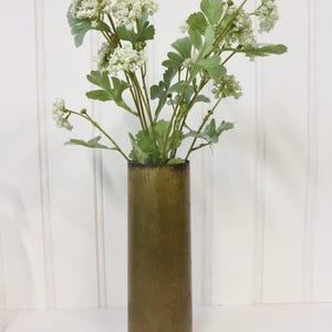 May include: A brass vase with a cylindrical shape, filled with white flowers and green leaves. The vase has a worn, aged look.