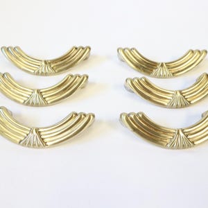 May include: Six gold-coloured, curved drawer pulls. Each pull features a fan-shaped design at the centre and a series of parallel ridges. The hardware is arranged on a white background, suggesting a home decor or furniture theme.