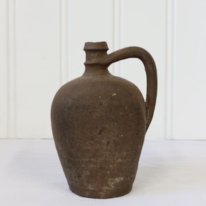 May include: A brown stoneware jug with a rounded body and a handle. The jug has a textured surface and a small opening at the top. The jug is likely an antique or vintage piece, suitable for rustic home decor.