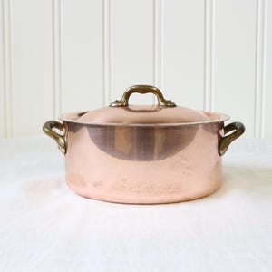 May include: A round copper pot with a matching lid, featuring two side handles and a central lid handle made of brass. The pot has a polished metallic finish with a warm, reddish-brown hue and a smooth, cylindrical body with a flat base.