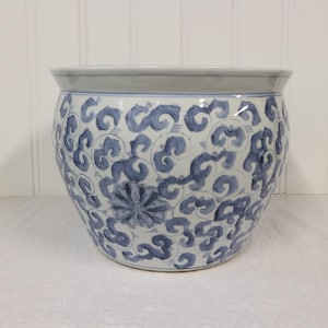 Pode incluir: Um vaso de cerâmica redondo com base branca e um padrão pintado à mão de flores e vinhas em azul. O recipiente possui um topo largo e aberto com uma borda lisa e alargada, além de uma base cónica, apresentando tons de azul e branco.
