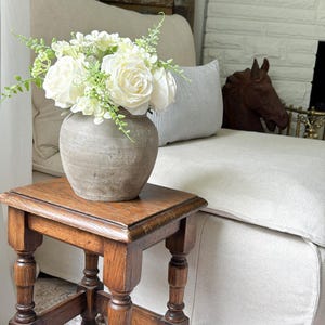 May include: A small, dark brown wooden stool with four turned legs and a square top. Resting on the stool is a round, textured grey ceramic vase filled with a bouquet of white artificial roses and small green leafy sprigs.