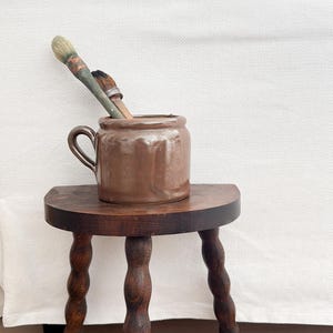 Peut inclure: Un pot en céramique marron avec une poignée et deux pinceaux repose sur un petit tabouret en bois foncé à trois pieds tournés. Le tabouret est sur un sol en bois clair, avec un fond blanc.