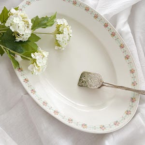 May include: An oval serving platter with a floral border in pink, yellow, and green. A silver serving utensil rests on the platter. White flowers and green leaves are placed near the platter on a white cloth.