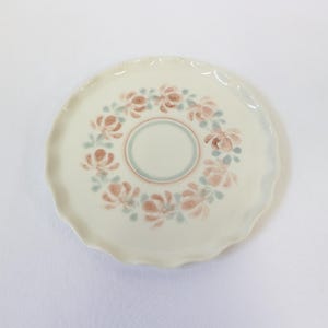 Peut inclure: Une assiette crème avec un bord festonné. L'assiette présente un motif floral dans les tons pêche et bleu autour du bord et un cercle central. L'assiette est probablement en céramique et convient pour servir des aliments.