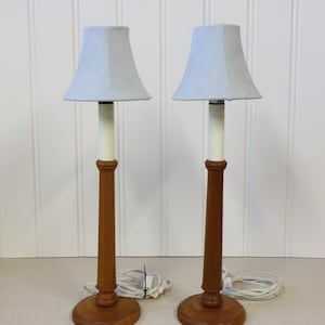 May include: A pair of table lamps featuring light blue fabric shades. The lamps have a wooden base and stem, with a white section connecting the shade. Each lamp has a white electrical cord.