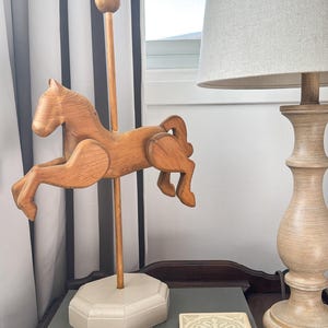 May include: A wooden carousel horse sculpture in a jumping pose. The horse is light brown and mounted on a wooden pole with a round top. It sits on a light grey, octagonal base. A lamp and a book are also in the image.