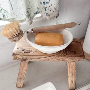 May include: A small, rustic wooden stool supports a white bowl containing a bar of soap and a wooden-handled brush. The soap is labelled "Oatmeal." A floral curtain hangs in the background. The stool is approximately 30 cm long.