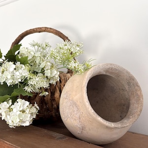 May include: A round, beige, weathered clay pot with a wide opening and a textured, matte finish. Next to the pot is a woven brown wicker basket filled with white hydrangea flowers and delicate green foliage, creating a natural arrangement.