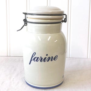 Peut inclure: Un pot de conservation en céramique de couleur crème avec l'inscription "farine" en écriture bleue sur le devant. Le pot est doté d'un couvercle assorti fixé par une fermeture à levier en métal noir et d'une fine bande bleue à la base.