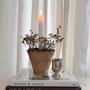 May include: A burning white candle stands on a stack of books, with a small terracotta pot of dried flowers in front. A silver cup is also on the books. The books are titled "WHITE & FADED" and "CREATE".