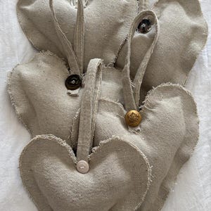 May include: A collection of five heart-shaped ornaments made from light beige, textured fabric with frayed edges. Each heart features a fabric hanging loop and is accented with a single, unique button in shades of dark brown, gold, or light pink.