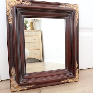 May include: A rectangular mirror with a dark brown wooden frame. The frame has ornate gold-coloured corner accents. The mirror is leaning against a white wall and a light brown wooden floor.