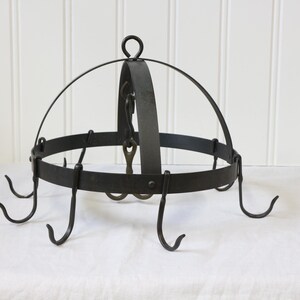 May include: A circular black metal pot rack featuring a central arched frame with a top hanging loop. The rack includes multiple curved hooks attached to the outer rim, designed for suspending kitchenware items in a hanging storage configuration.