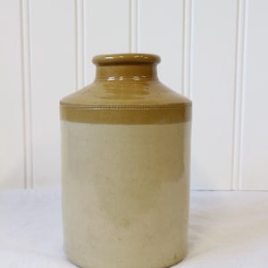 May include: A cylindrical stoneware crock featuring a two-tone glaze design. The upper portion and rim are finished in a glossy brown glaze, while the lower body is a matte cream colour. A subtle decorative band with a dotted texture separates the colours.