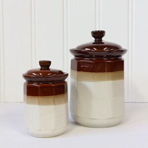 May include: Two ceramic storage jars with lids. The jars are brown, tan, and white with a glossy finish. The smaller jar is about 15 cm tall, and the larger jar is about 23 cm tall. The jars are likely for kitchen storage.