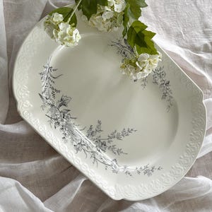 May include: An off-white, oval serving platter with a delicate floral design in grey. The platter has a scalloped edge and is adorned with a sprig of white flowers. The background is a white, textured fabric.