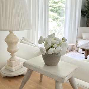 May include: A small, round, speckled grey ceramic vase filled with white ranunculus flowers, resting on a white, weathered wooden stool with four splayed legs, positioned next to a white table lamp with a pleated shade on a wooden surface.