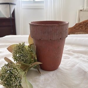 May include: A terracotta-coloured plant pot with a vertical ribbed texture along the exterior and a wider, reinforced rim at the top. The pot is positioned next to a decorative stem featuring small green flower buds and light green leaves.