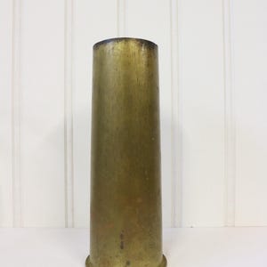 May include: A brass shell casing vase. The vase is cylindrical in shape with a slightly flared top. The brass has a worn, aged appearance.