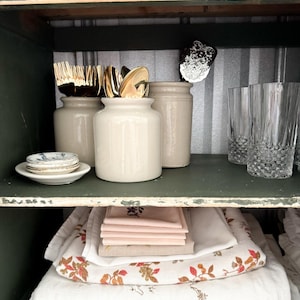 Peut inclure: Une armoire en bois blanc avec deux étagères. L'étagère supérieure contient trois pots en céramique blanche avec des couverts en argent doré à l'intérieur. L'étagère inférieure contient une pile de serviettes en lin blanc avec un motif floral.