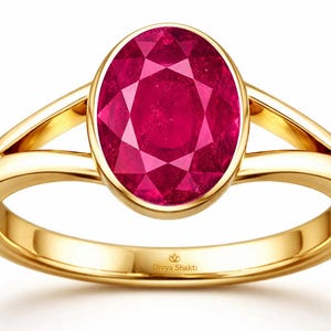 May include: A gold ring featuring a large, oval-cut, deep pink gemstone set in a smooth bezel setting. The band has a split-shank design that connects to the central stone setting. The interior of the band is engraved with the text Divya Shakti.