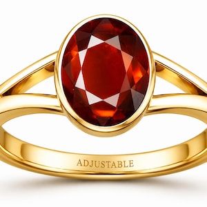 May include: A gold ring featuring a large, oval-cut, deep red gemstone set in a smooth bezel. The band has a split-shank design and the word ADJUSTABLE is clearly engraved on the inner surface of the gold band, indicating its functional feature.