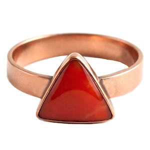 May include: A copper-colored ring with a triangular, red gemstone centerpiece. The ring band is a simple, rounded design, and the gemstone is set in a bezel setting. The ring is likely a fashion accessory.