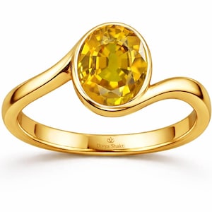 Könnte beinhalten: Ein Goldring mit einem zentralen, oval geschliffenen gelben Edelstein in einer glatten Fassung. Die Ringschiene hat ein geschwungenes Design, das den Stein umschließt. Innen ist der Ring mit Divya Shakti und einem Lotus-Symbol graviert.
