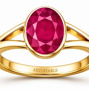 May include: A gold ring featuring a large, oval-cut, deep pinkish-red gemstone set in a smooth bezel. The band has a split-shank design and the word ADJUSTABLE is engraved on the inner surface of the gold band, clearly visible in the center.