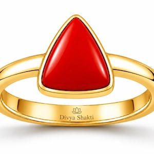 May include: A gold ring featuring a smooth, triangular red stone set in a polished gold bezel. The inner band of the ring is engraved with the text Divya Shakti and a small lotus flower icon, showcasing a clean, reflective metallic finish throughout.