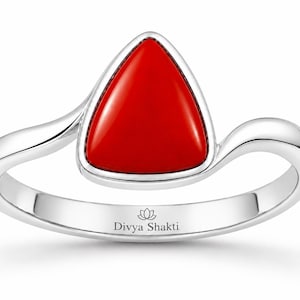 May include: A silver ring featuring a smooth, triangular red stone set in a polished metal band. The band has a curved, asymmetrical design and includes the text Divya Shakti with a small lotus flower icon engraved on the inner surface of the ring.