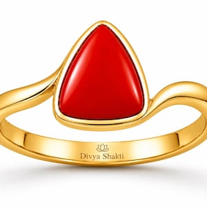 Op de afbeelding: Een gouden ring met een gladde, driehoekige rode steen gezet in een gepolijste gouden band. De band heeft een gebogen ontwerp in bypass-stijl. De tekst Divya Shakti en een klein lotusicoon zijn gegraveerd op de binnenzijde van de ringband.