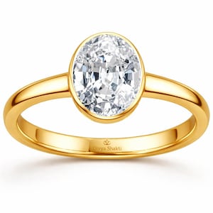 May include: A gold ring featuring an oval-cut diamond. The band is polished gold, and the ring is set against a white backdrop. The text "Divya Shakti" is engraved on the band. A classic piece of jewellery.
