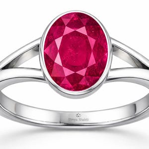 Op de afbeelding: Een zilveren ring met een grote, ovaal geslepen, diep roze-rode edelsteen in een gladde zetting. De ringband heeft een gespleten ontwerp dat aansluit op de centrale zetting. De binnenzijde is gegraveerd met de tekst Divya Shakti.