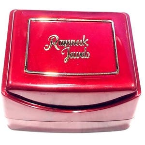 May include: A glossy, deep red rectangular jewellery box with a hinged lid. The centre of the lid features a gold-toned rectangular border surrounding the gold-coloured embossed text that reads Ramneck Jewels in a cursive script font.