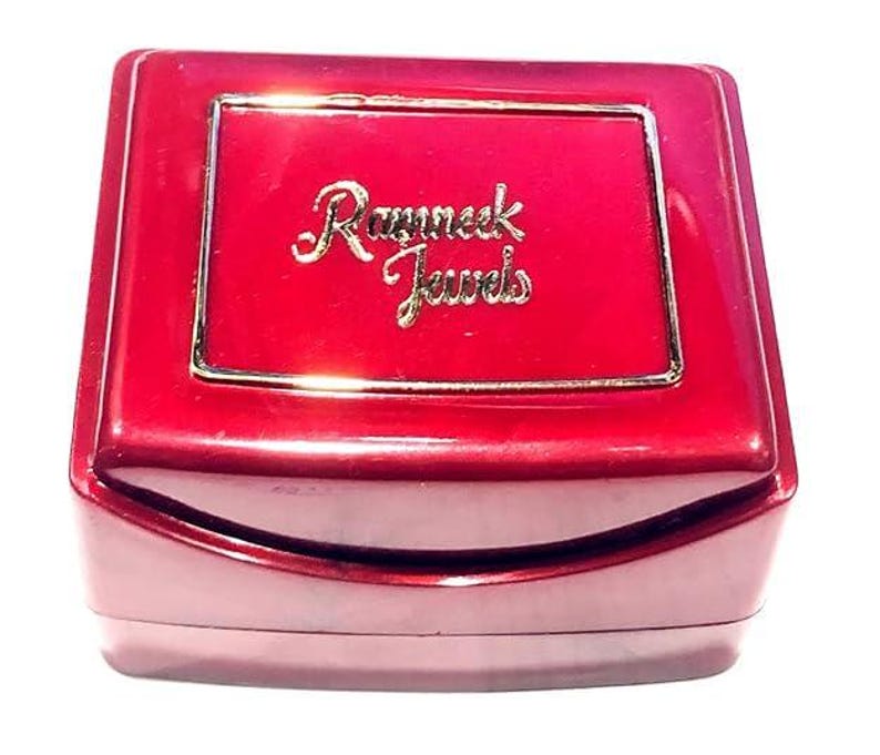 May include: A glossy, deep red rectangular jewellery box with a hinged lid. The centre of the lid features a gold-toned rectangular border surrounding the gold-coloured embossed text that reads Ramneck Jewels in a cursive script font.