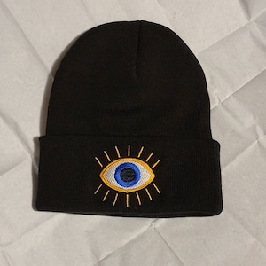 May include: Black beanie with an embroidered blue eye with yellow rays. The eye is a traditional evil eye symbol.