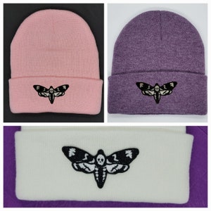 May include: Three knit beanies with a black and white moth graphic. The beanies are pink, purple, and white.