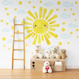 May include: A nursery room with a white wall decorated with a yellow sun with a smiling face, blue clouds, and yellow dots. There is a wooden ladder, a wooden storage box, and five teddy bears in the room.