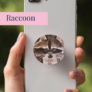 May include: A white phone case with a round pop socket featuring a watercolor illustration of a raccoon's face. The text "Raccoon" is written in pink above the pop socket. The text "Simple Designs by Elizabeth" is written in white on a pink background with floral accents.