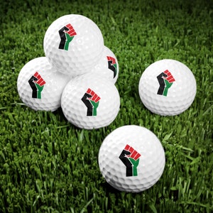 May include: Six white golf balls with a black fist design and red, green, and black accents. The golf balls are arranged in a semi-circle on a green surface.