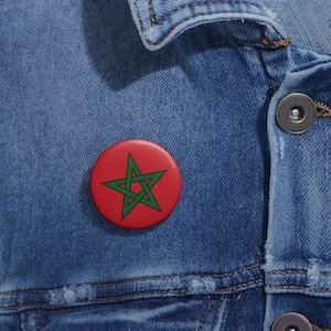 May include: A round red button with a green star emblem, likely a national symbol, is pinned to a blue denim jacket. The button's design is simple and bold, with a clear contrast between the red background and the green star.