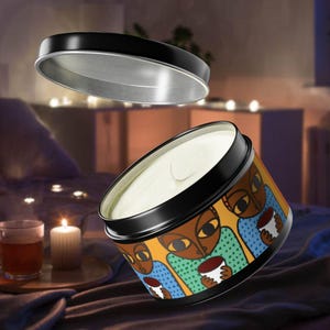 Puede incluir: Una vela en un recipiente de hojalata con una ilustración colorida de tres personas sosteniendo tazas. La vela es blanca y la hojalata es negra con una tapa plateada.